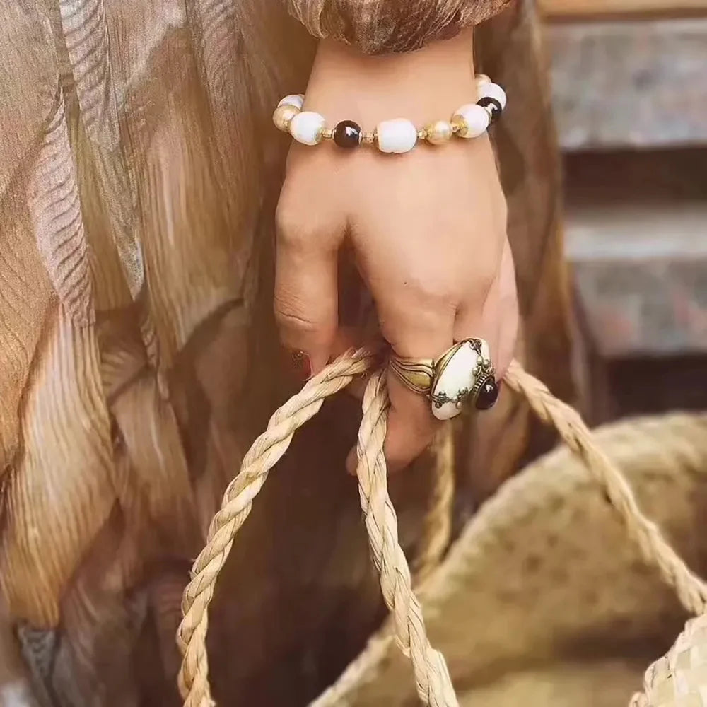 Bague ethnique avec une pierre blanche et une petite pierre noire, montée sur un anneau doré, portée par une main tenant un bracelet de perles blanches et noires et une anse de panier en osier.