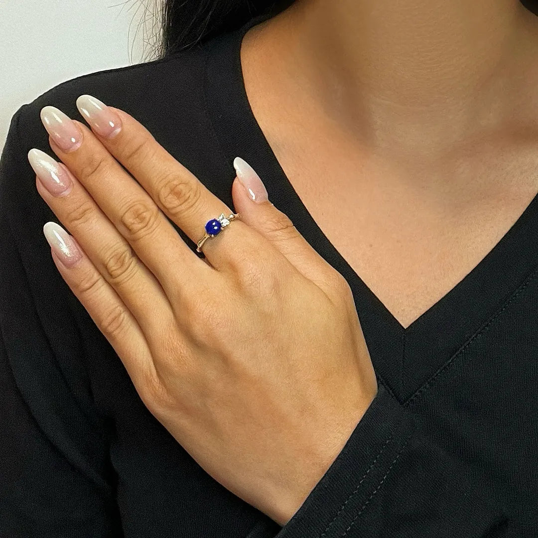 Bague en argent avec un lapis lazuli bleu profond au centre et deux petites pierres blanches de chaque côté, portée à l'annulaire d'une main aux ongles vernis.