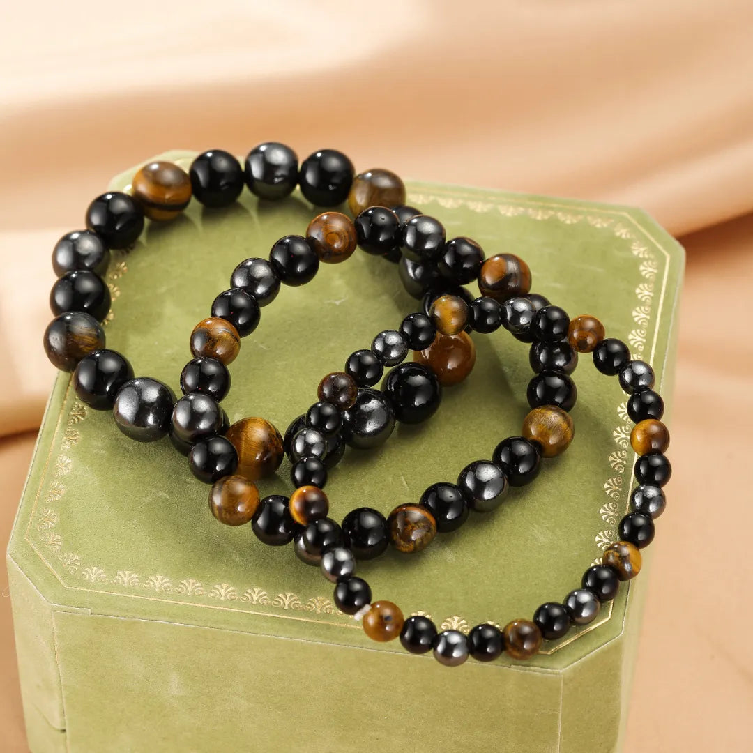 Trois bracelets faits de perles d'œil de tigre et d'hématite disposés sur une boîte verte ornée, symbolisant la force protectrice.