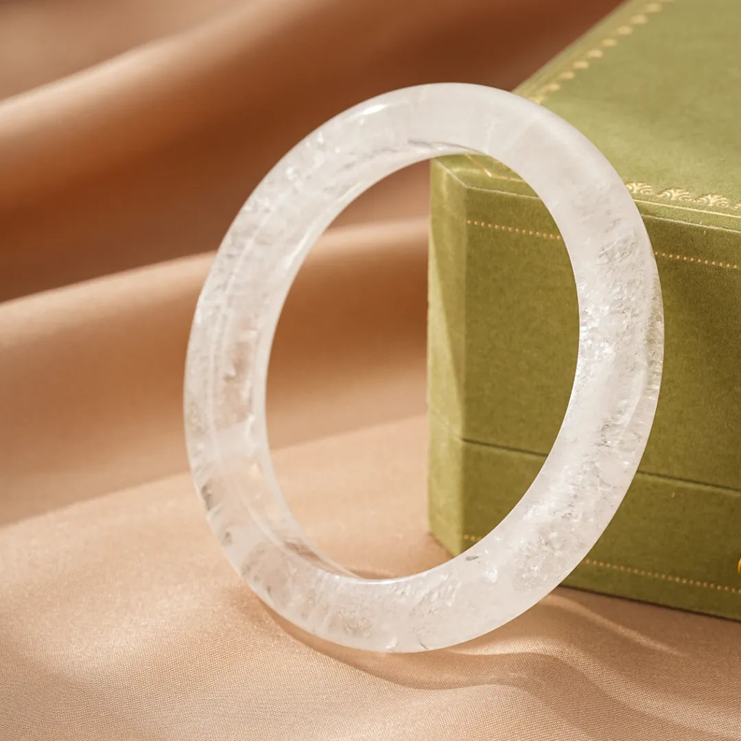 Bracelet jonc en cristal de roche naturel, bijou "Pureté Lumineuse" transparent et élégant, posé sur un fond satiné près d'une boîte verte.