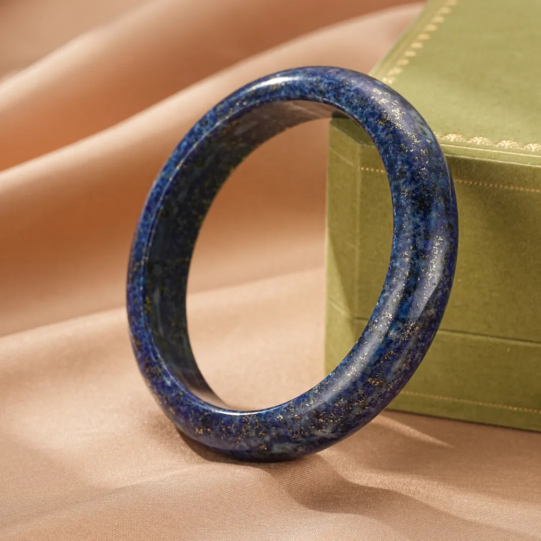 Bracelet jonc en lapis lazuli bleu nuit avec inclusions dorées, sur un fond satiné beige et une boîte cadeau verte.