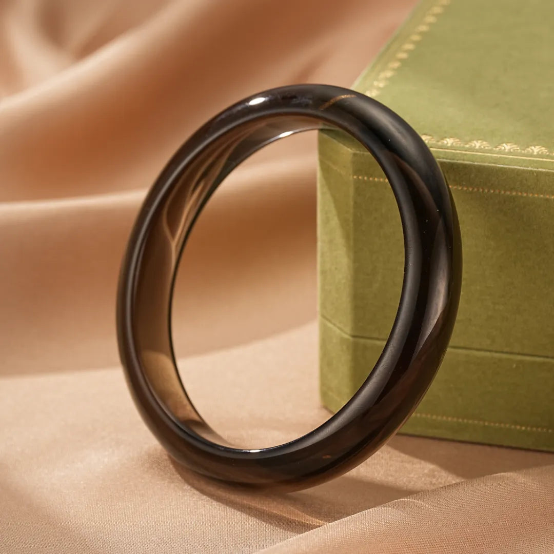 Bracelet jonc en obsidienne "Mystère Profond" de couleur noire, posé sur un tissu satiné beige avec une boîte cadeau verte en arrière-plan.