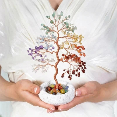 Arbre de Vie 7 Chakras en pierres naturelles colorées monté sur un tronc en cuivre, présenté dans un pot blanc texturé, tenu par des mains sur fond blanc.
