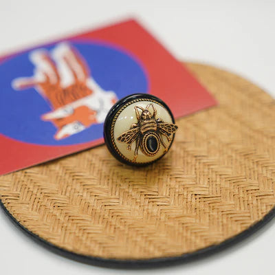 Bague Agate Noire "Reine Noire" avec un motif d'abeille dorée sur fond crème, entourée d'un anneau noir, posée sur un napperon en osier avec une carte en arrière-plan.
