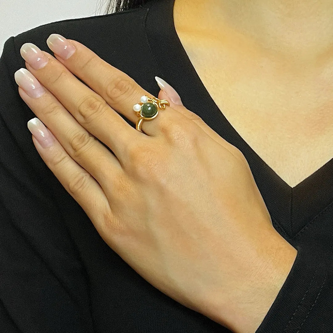 Bague en or 14 carats avec une pierre verte centrale et deux petites perles blanches, ornée d'un design rappelant un chaton joyeux. La bague est portée sur l'annulaire d'une main aux ongles soignés.