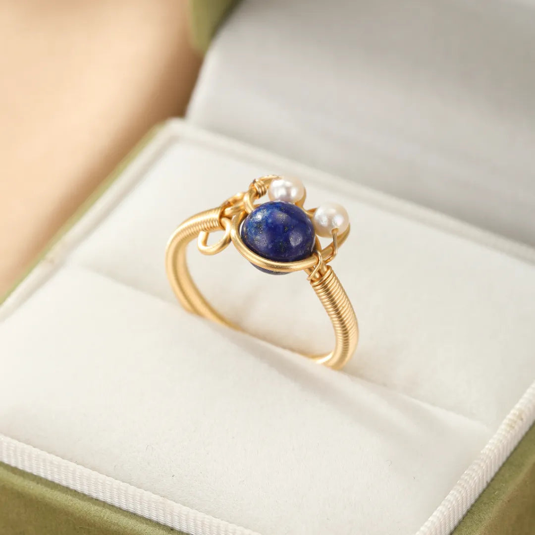 Bague en filigrane doré or 14K sertie d'une pierre bleue lapis-lazuli et de deux petites perles blanches, posée dans une boîte à bijoux verte.
