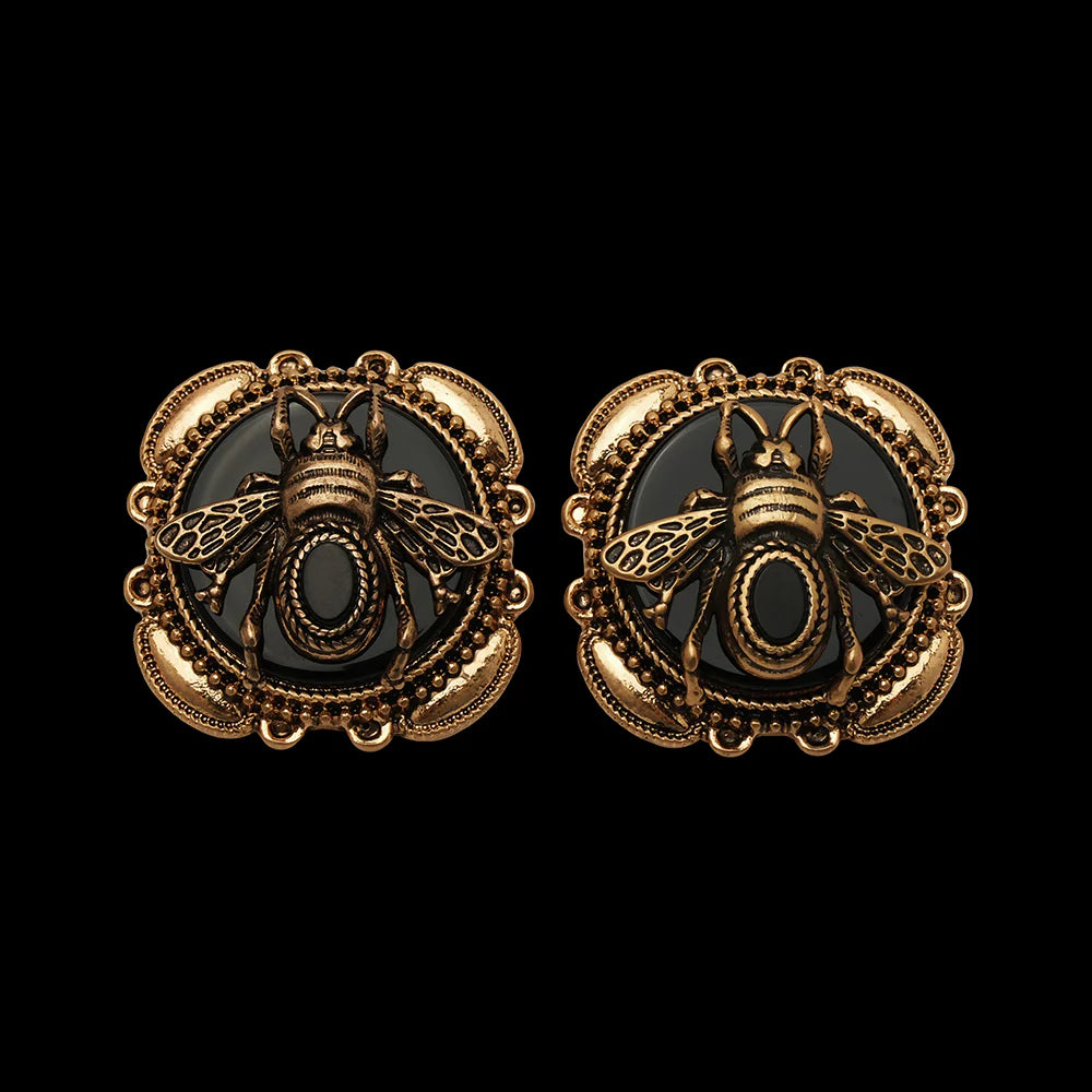 Paire de boucles d'oreilles plaquées or avec une abeille stylisée sur fond d'agate noire, bordées d'un cadre orné de motifs délicats.