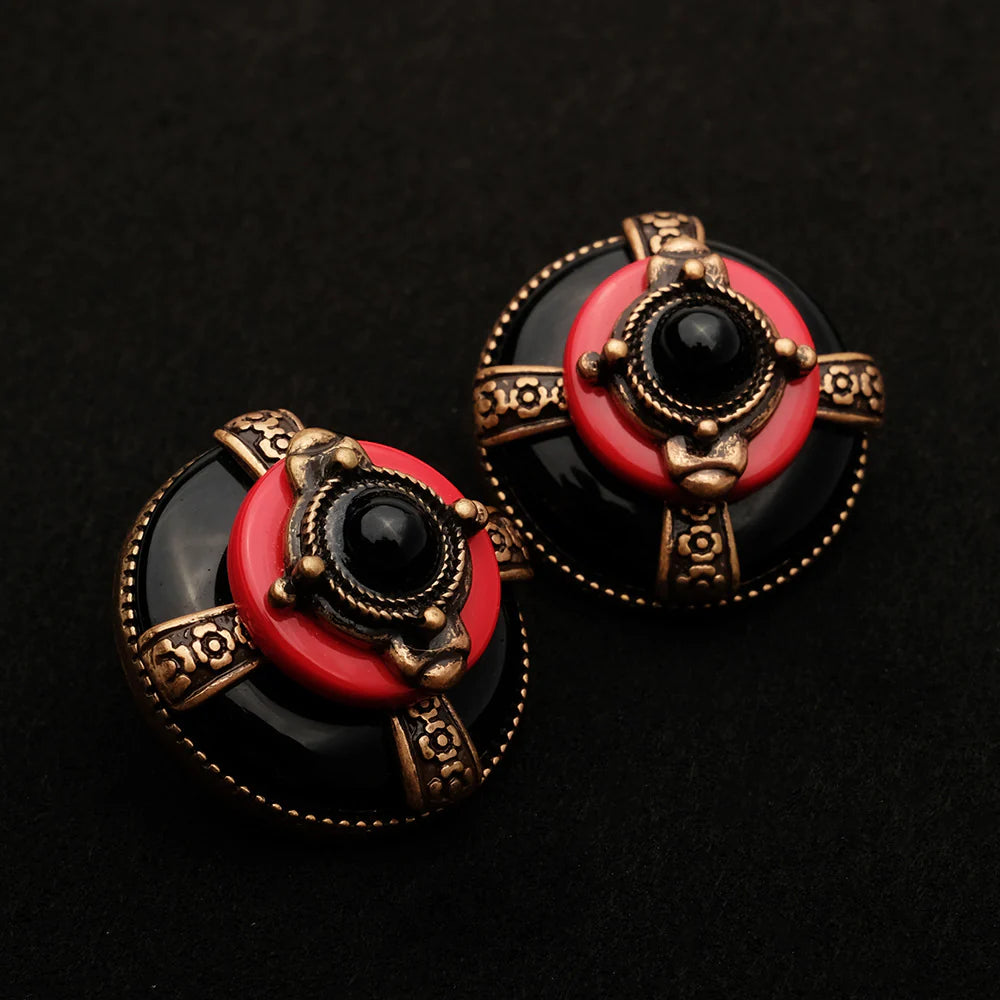 Boucles d'oreilles rondes avec une pierre noire au centre, ornées de détails dorés et d'un cercle rouge, évoquant un style vintage.
