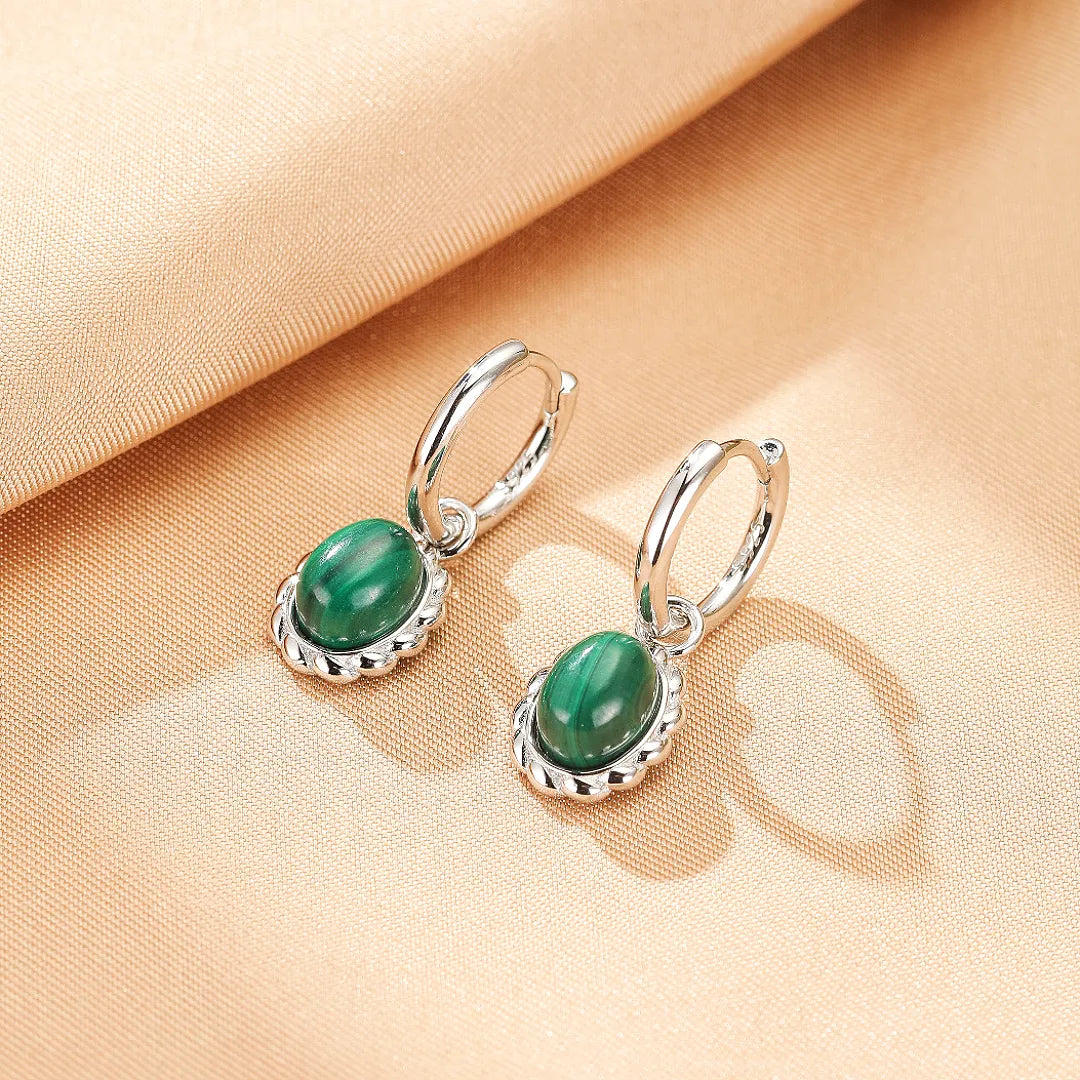 Paire de boucles d'oreilles en argent avec une pierre de malachite verte ovale et une monture finement ciselée, sur un fond texturé beige.