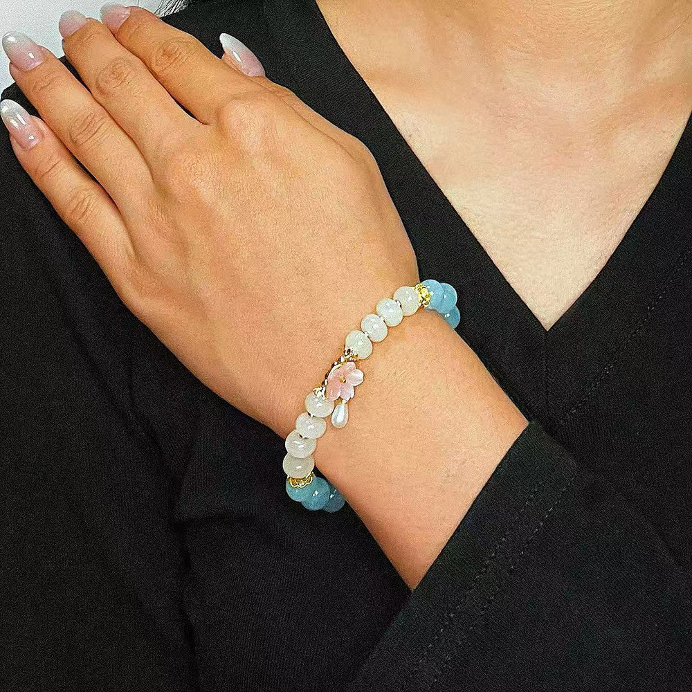 Bracelet "Pureté de Matin" en aigue-marine et agate avec un pendentif fleur rose et une perle blanche, porté au poignet d'une femme.