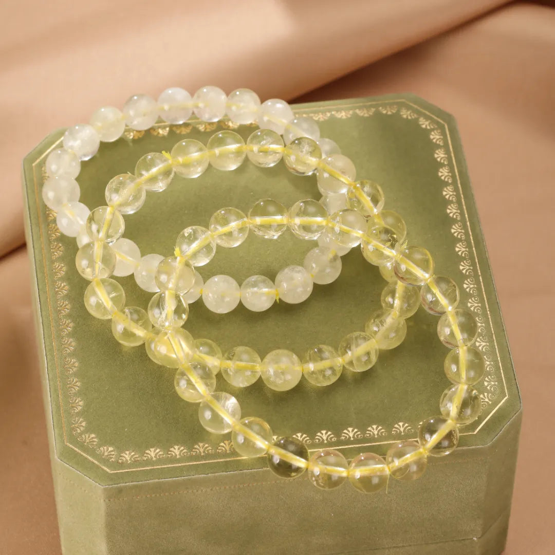 Trois bracelets en citrine naturelle sur une boîte cadeau verte ornée de motifs dorés. Les perles sont rondes et translucides, de couleur jaune pâle à dorée.