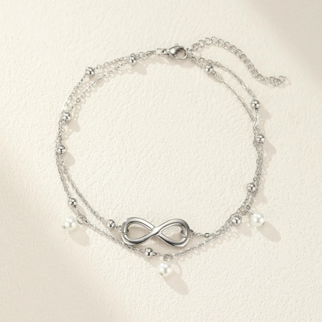 Bracelet de cheville double chaîne argenté orné d'un symbole infini et de perles blanches nacrées.