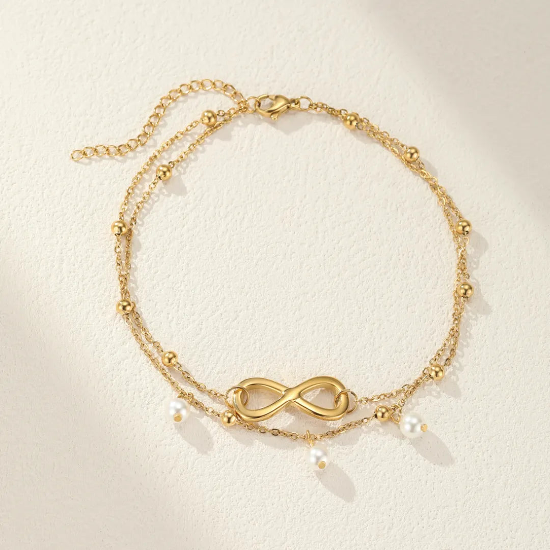 Bracelet de cheville double chaîne doré avec un symbole infini central et des perles nacrées suspendues, style bohème chic.