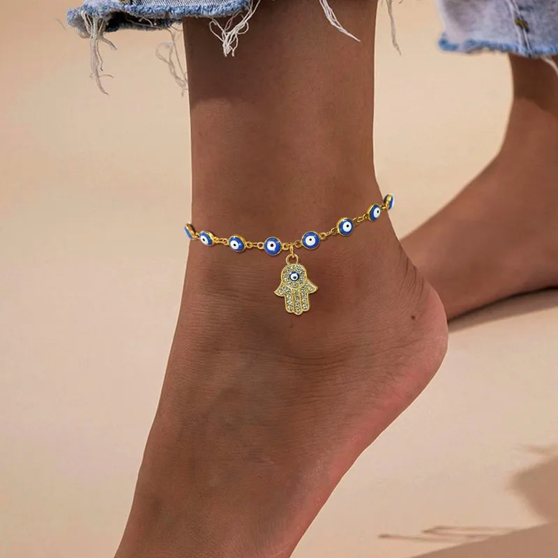 Bracelet de cheville doré orné de multiples petits yeux bleus et blancs, avec une breloque centrale en forme de main de Fatma (Khamsa) incrustée de strass.