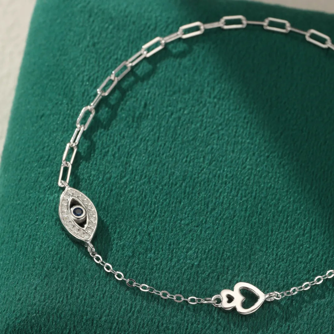 Bracelet de cheville en argent 925 orné d'un motif œil stylisé serti de petites pierres scintillantes et d'un saphir bleu au centre, avec une délicate chaîne à maillons et un charmant motif de cœurs entrelacés.
