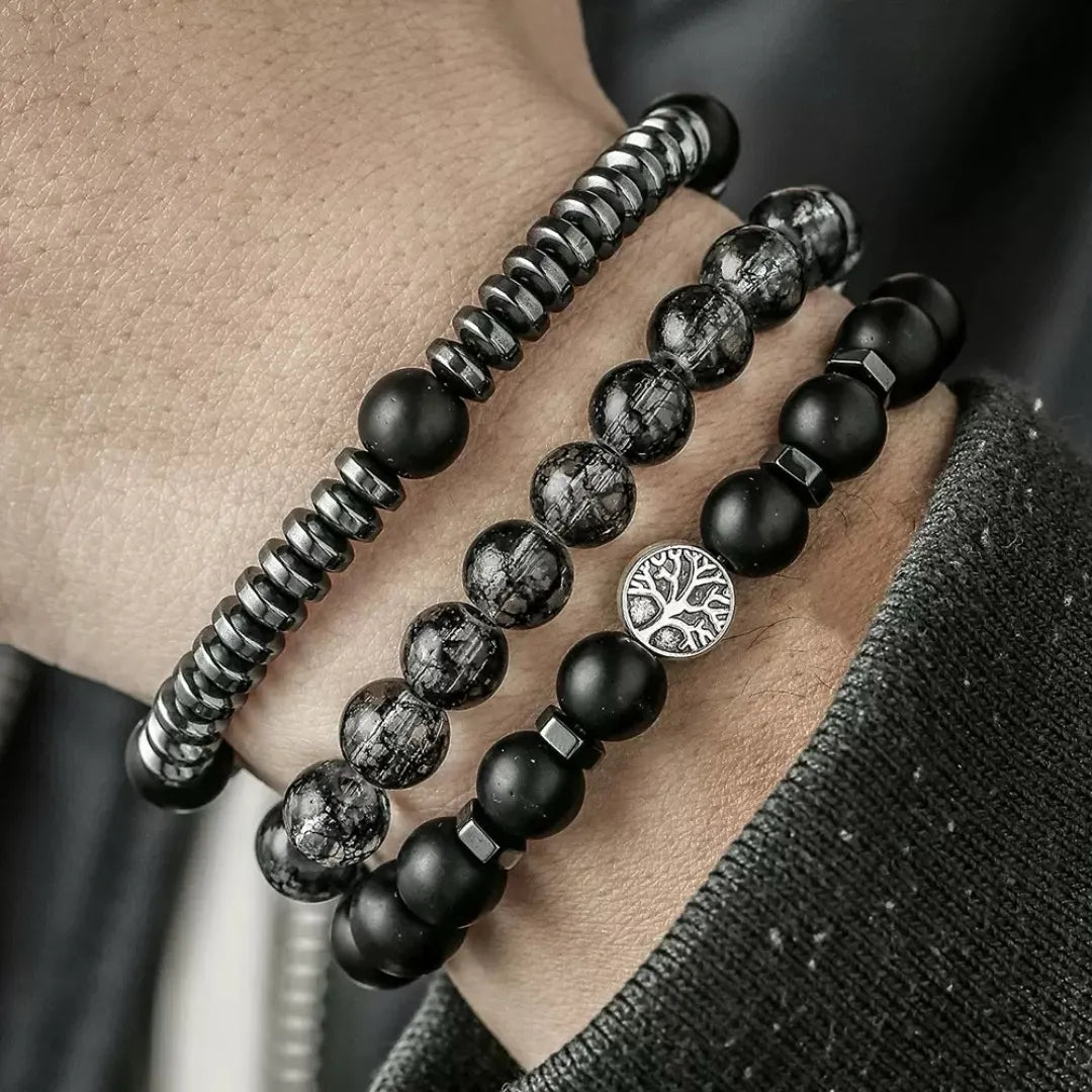 Trois bracelets pour hommes empilés sur un poignet : un bracelet en hématite striée, un bracelet en obsidienne noire avec un pendentif arbre de vie argenté et un bracelet en perles d'obsidienne intercalées de petits cubes métalliques.