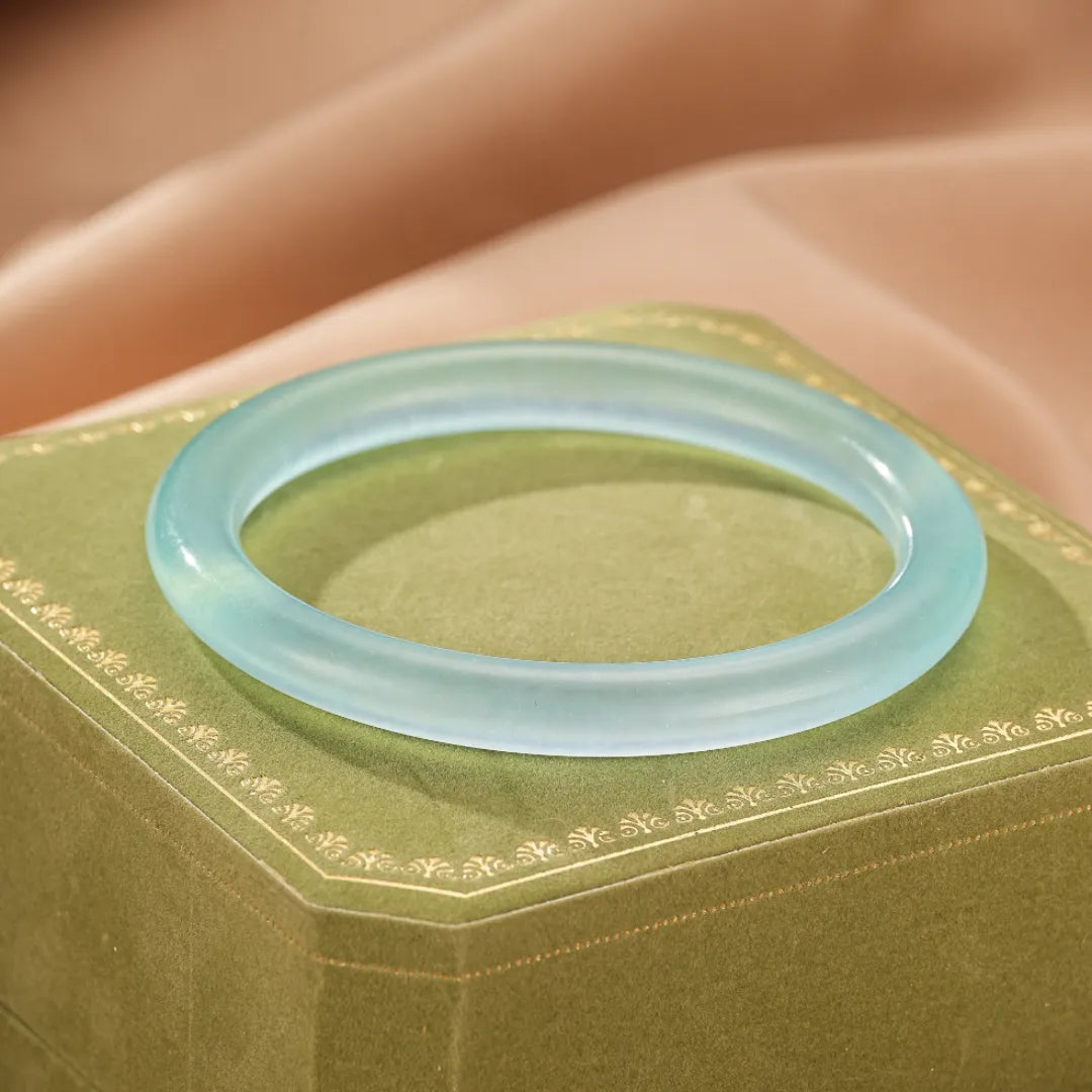 Bracelet jonc en aigue-marine translucide, posé sur un écrin vert orné de motifs dorés. La pierre a une teinte bleu clair évoquant la douceur de la mer.
