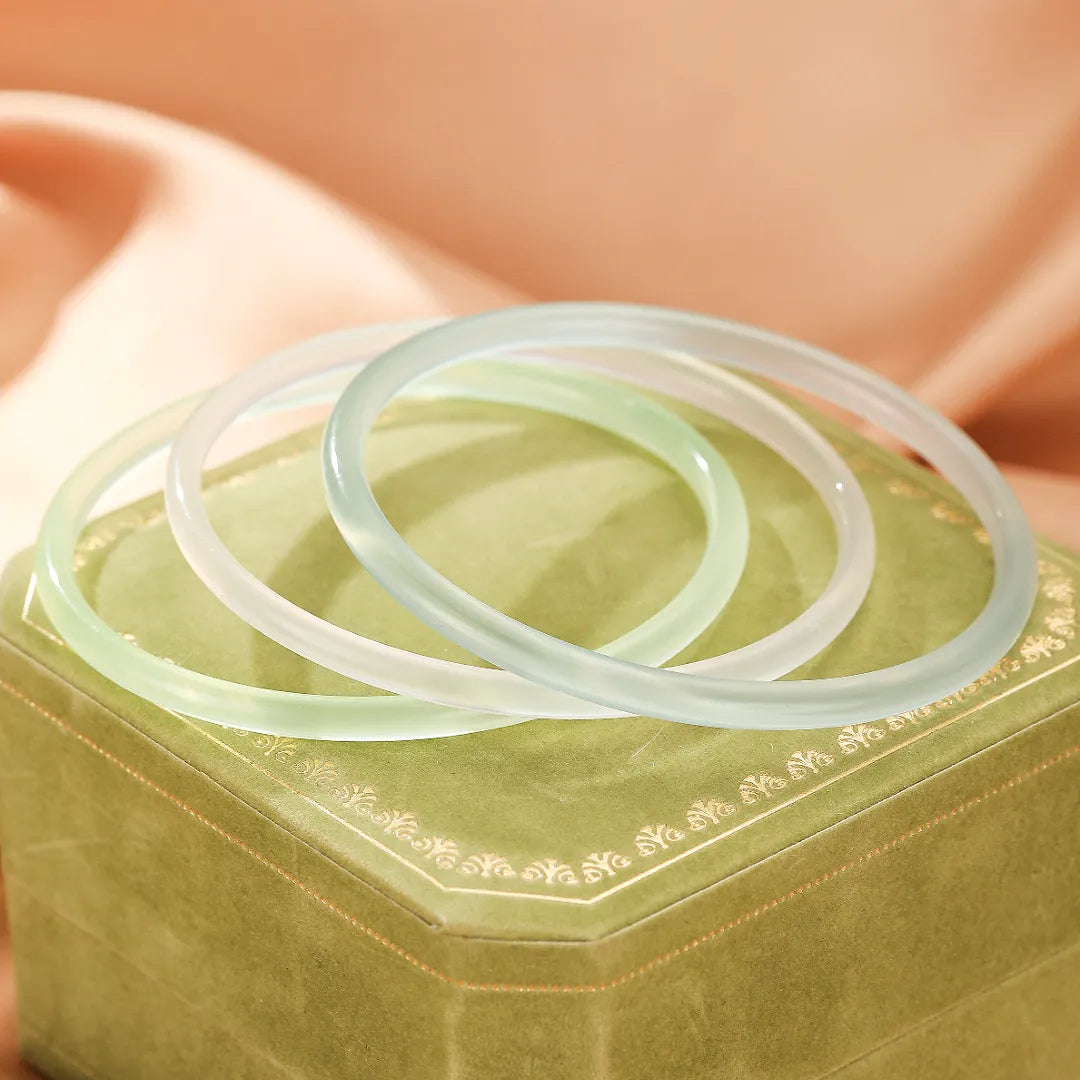 Trois bracelets jonc en calcédoine aux teintes pastel douces, posés sur une boîte de présentation verte ornée, évoquant la fraîcheur aquatique.