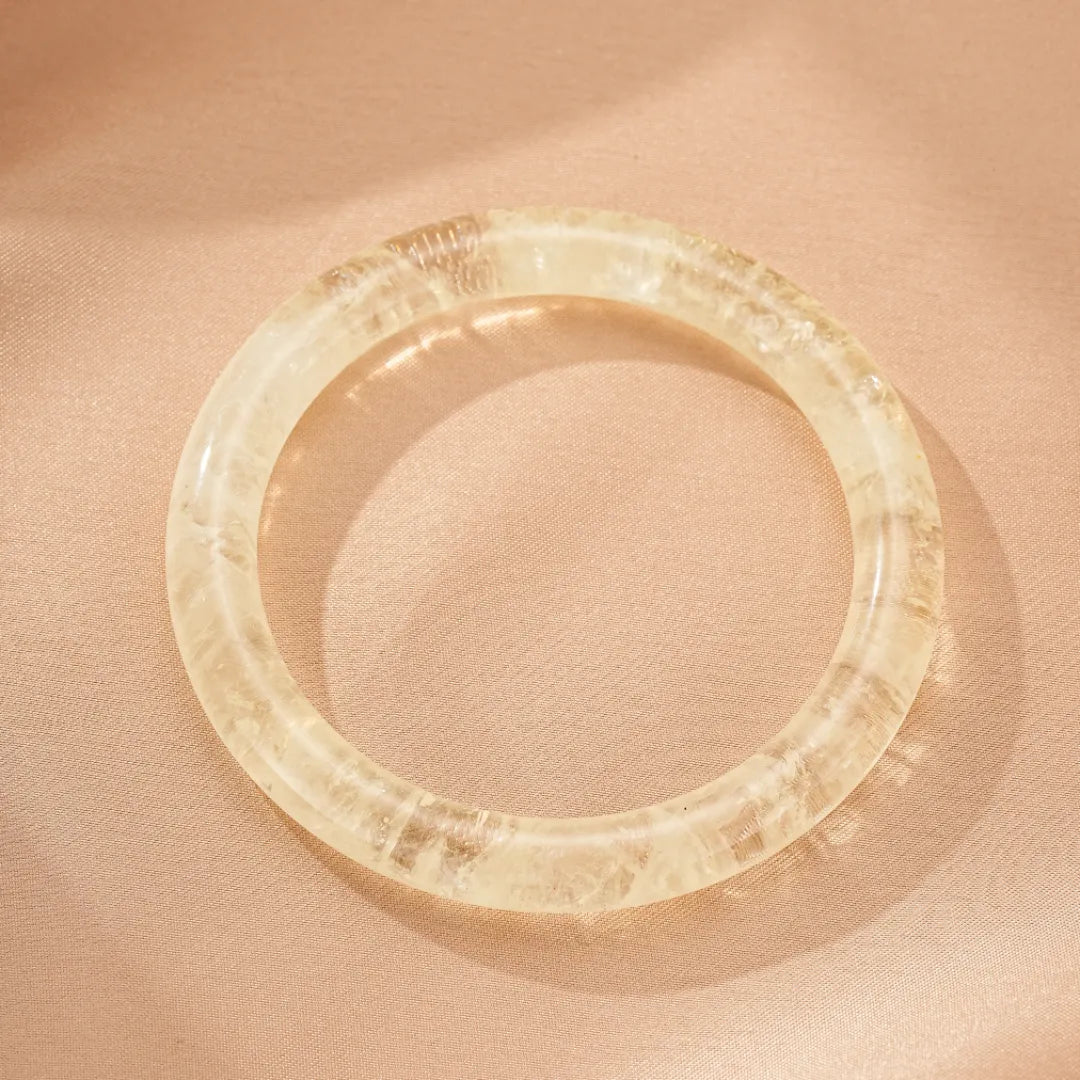 Bracelet jonc en citrine naturelle transparente "Reflets de Confiance" posé sur un tissu satiné couleur pêche. La pierre présente des inclusions et des reflets lumineux.