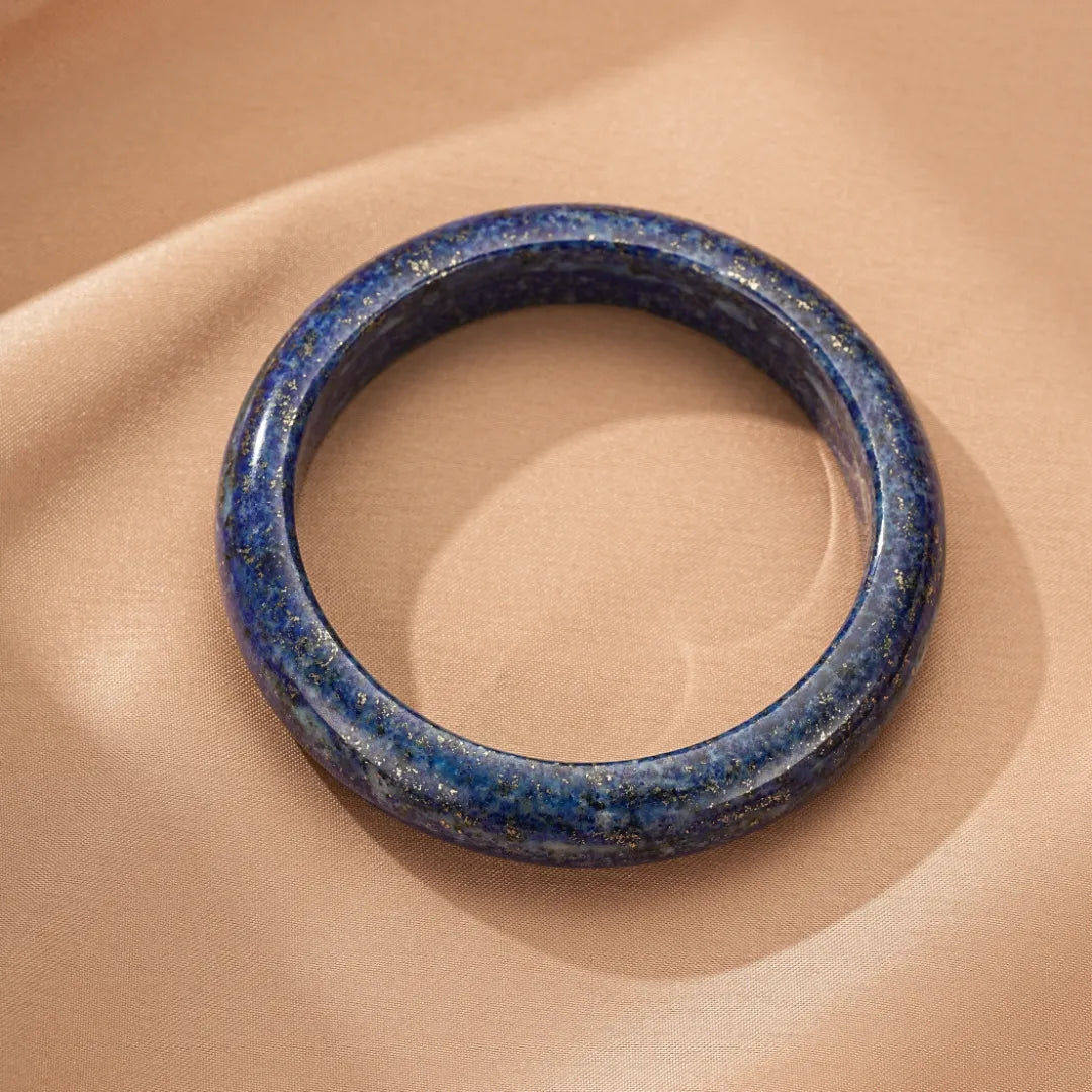 Bracelet jonc en lapis lazuli bleu profond avec des inclusions dorées, posé sur un tissu satiné beige.