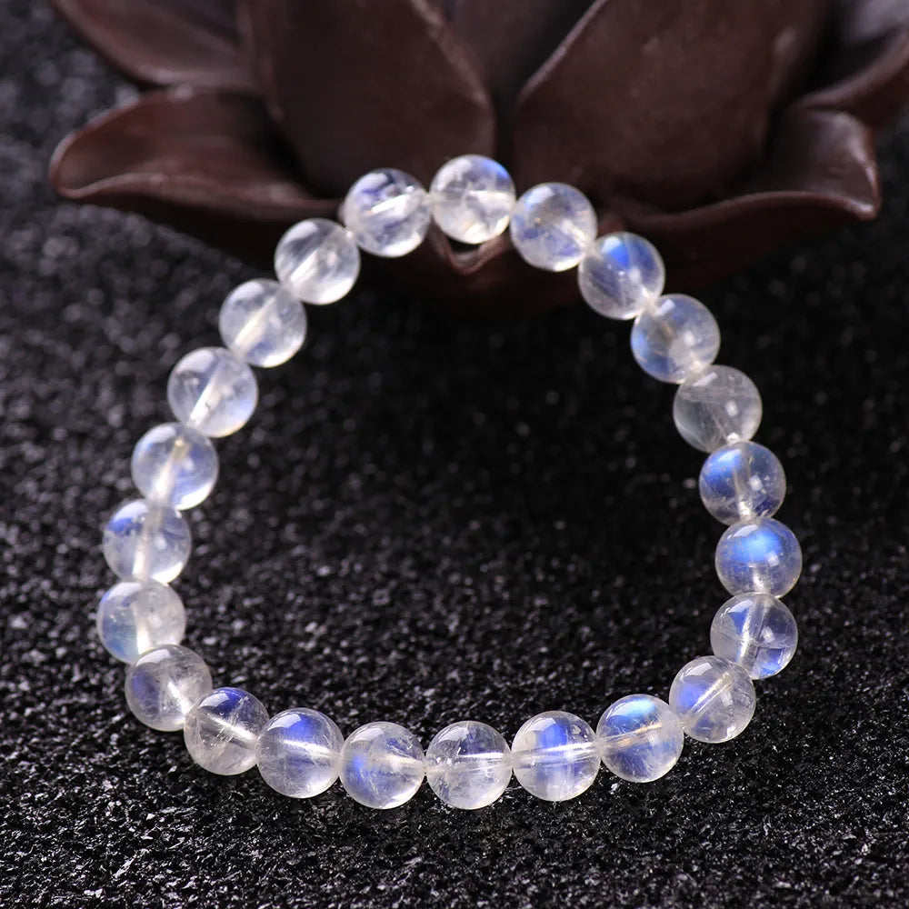 Bracelet en pierres de lune naturelles "Rêve Doux" avec des perles rondes translucides aux reflets bleutés et blancs, posé sur un fond noir texturé.