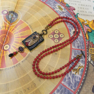 Collier "Beauté Naturelle" avec pendentif rectangulaire noir orné d'une cigale dorée et d'une pierre rouge, sur un collier de perles rondes rouges.