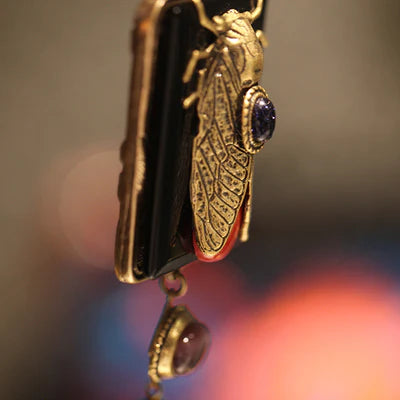 Collier avec une tête d'agrion dorée, un corps orné d'une pierre bleue et des ailes détaillées sur fond noir, avec une gouttelette suspendue.