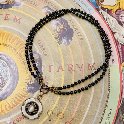 Collier Agate Noire "Pureté Mystique" composé de deux rangs de perles noires et d'un pendentif rond blanc orné d'une pierre centrale et de détails dorés, sur fond de carte astronomique.