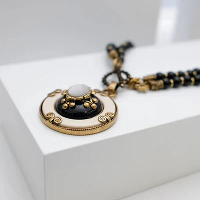 Collier Agate Noire "Pureté Mystique" avec pendentif rond en émail blanc et noir, orné de perles dorées et d'une pierre blanche centrale, sur une chaîne de perles noires.