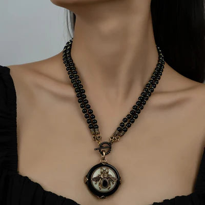 Collier Agate Noire "Reine Noire" avec pendentif rond noir et blanc orné d'une abeille dorée et d'une pierre centrale noire, monté sur deux rangs de perles noires.