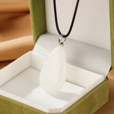 Collier avec un pendentif en forme de goutte en cristal de roche blanc, suspendu à un cordon noir, présenté dans un écrin vert.