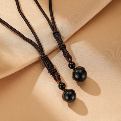 Gros plan sur deux pendentifs de collier en obsidienne en forme de boule, fixés sur des cordons marron tressés, sur un fond de tissu satiné couleur crème.