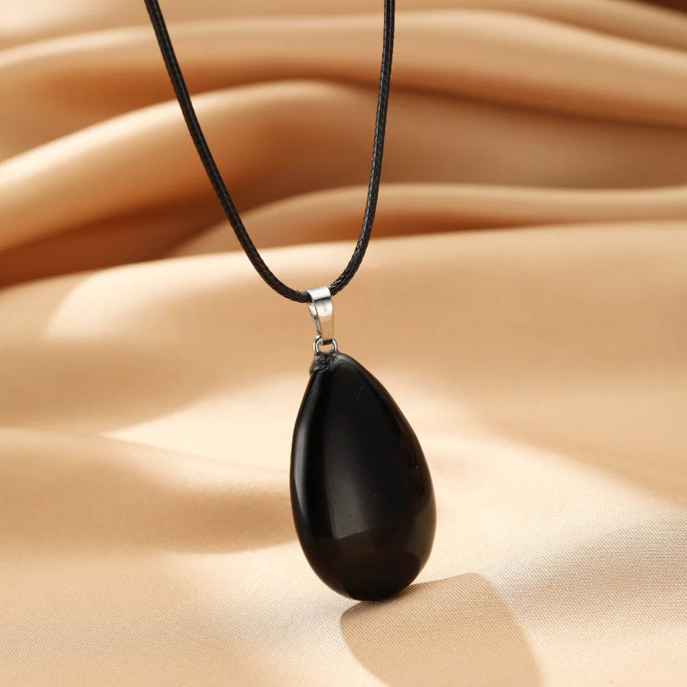 Collier pendentif en forme de goutte d'obsidienne noire polie, monté sur un cordon noir, sur un fond satiné beige.