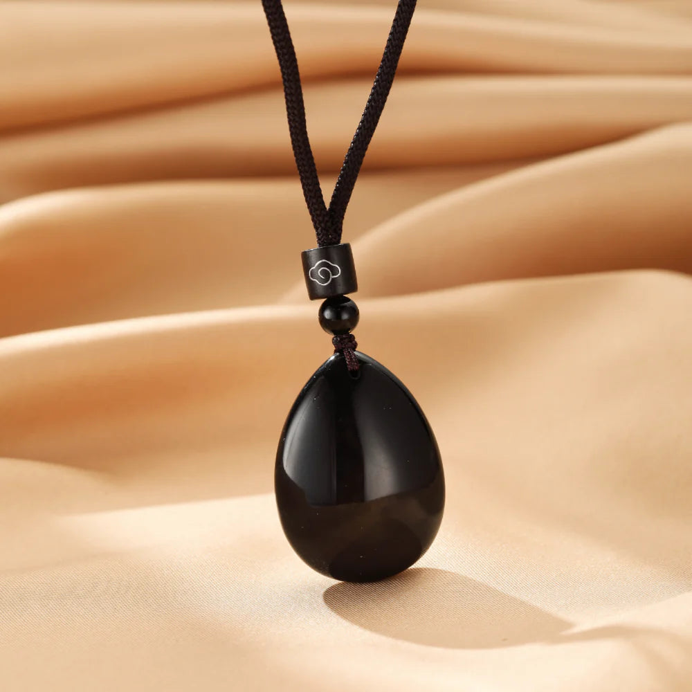 Collier avec pendentif en forme d'œuf en obsidienne noire lisse, suspendu à un cordon noir avec une perle et un cube décoré d'un motif de nuage stylisé.