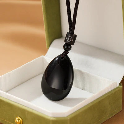 Collier avec un pendentif en forme de goutte d'obsidienne noire polie, suspendu à un cordon noir réglable avec une perle et un petit élément décoratif gravé d'un nuage.